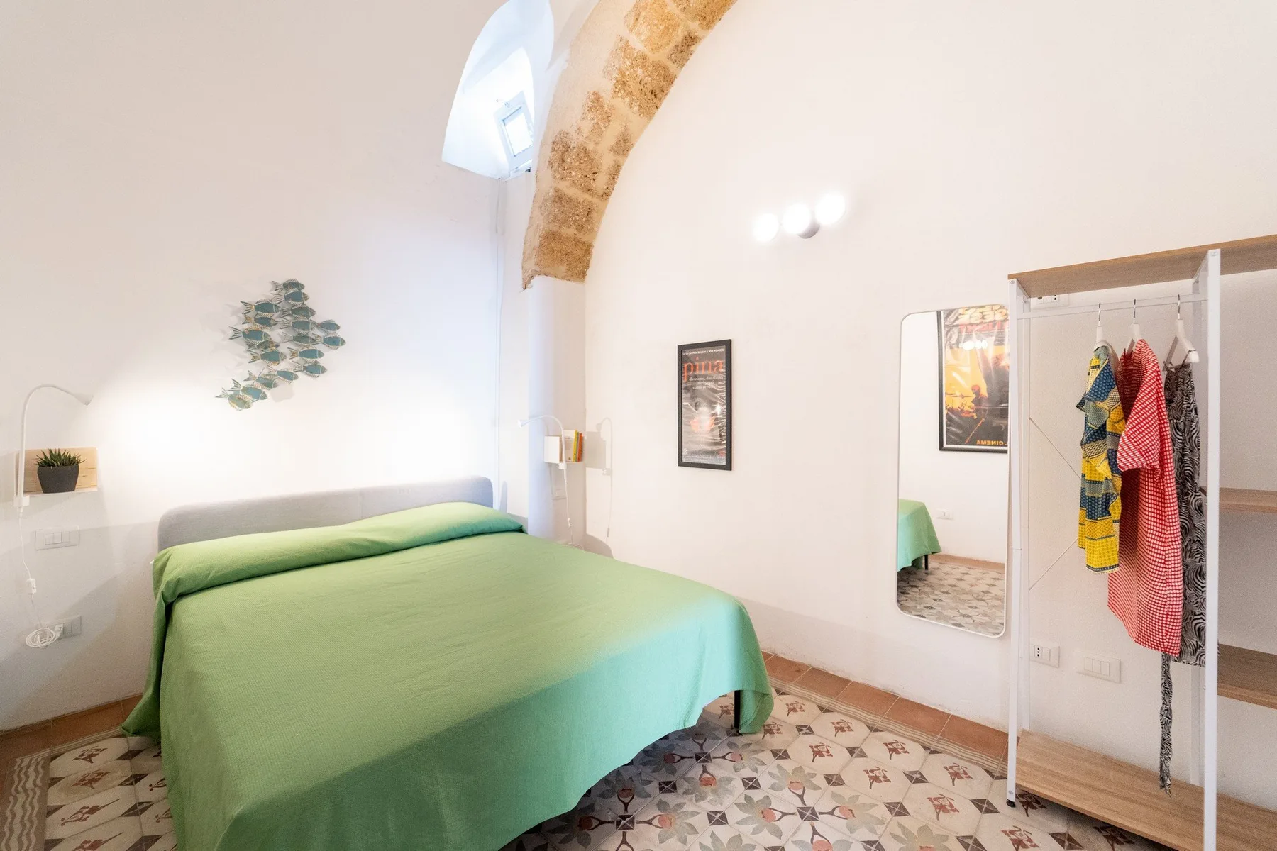 A bedroom with green bedding, a wall mirror, and an open clothes rack.