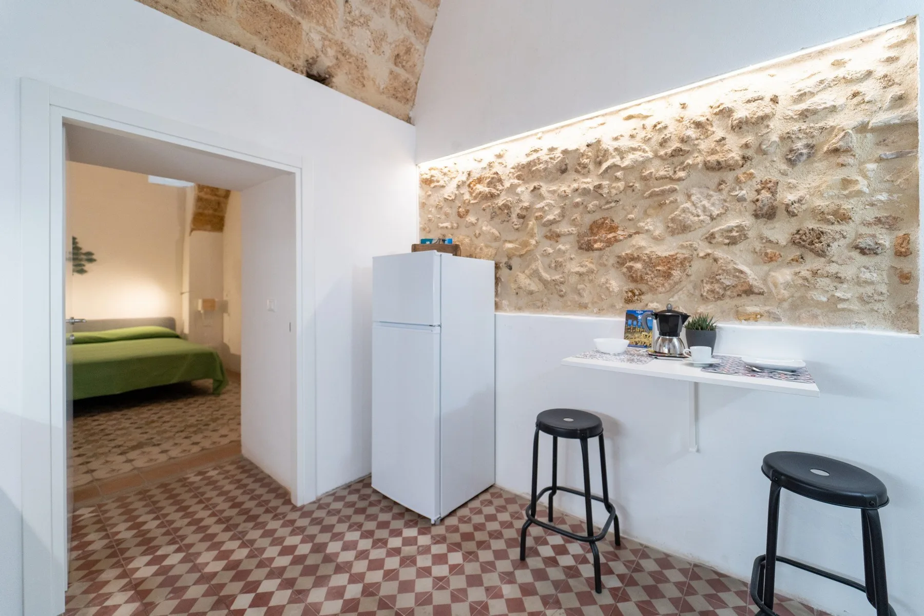 A bright kitchenette with a refrigerator and counter stools beside a stone wall.
