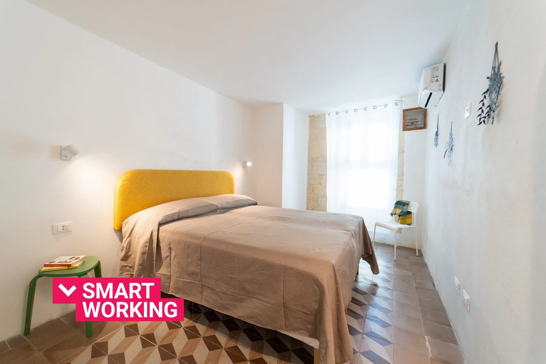 A bedroom with a double bed, yellow headboard, and bright window.