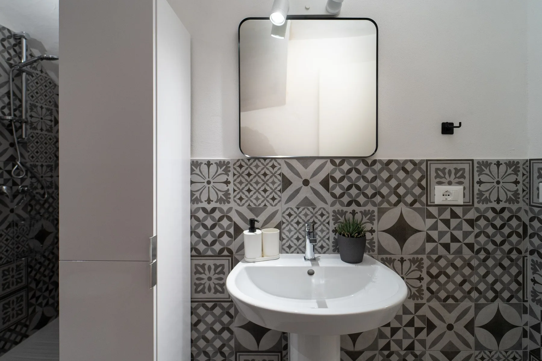 A bathroom sink sits beneath a mirror, with patterned tiles on the wall.