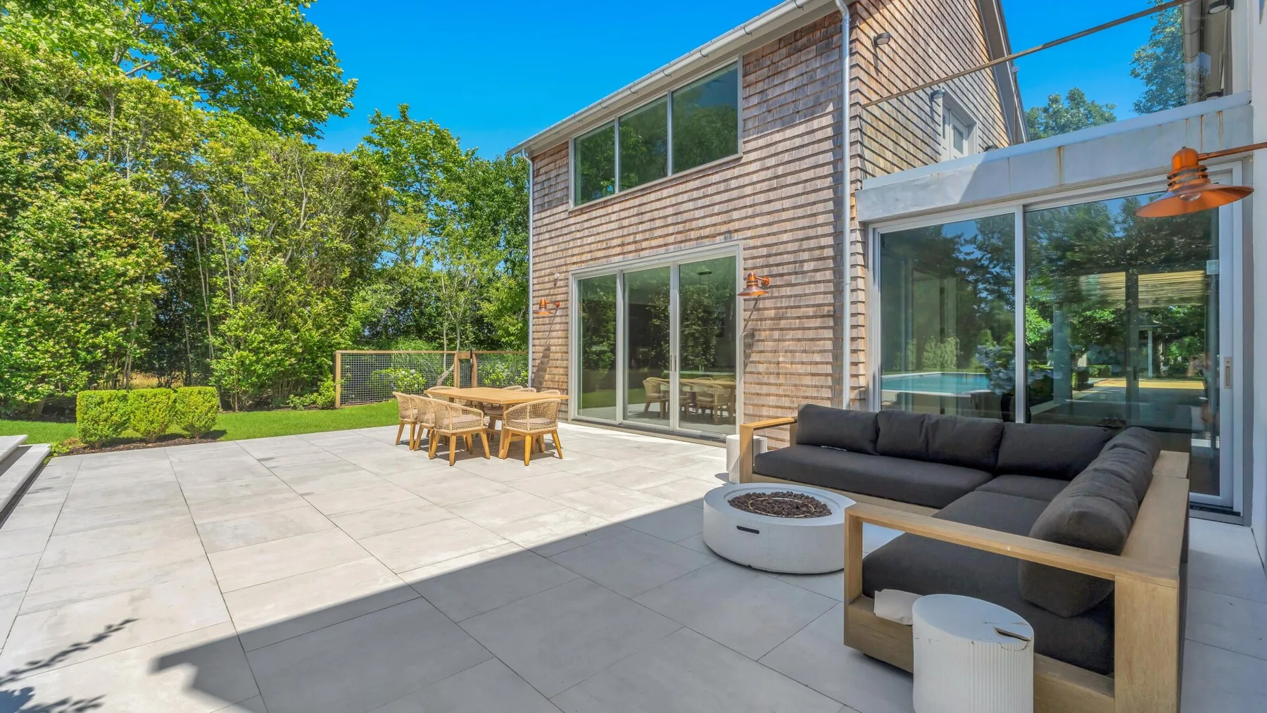 Una casa moderna da a un patio pavimentado con asientos al aire libre y árboles alrededor.