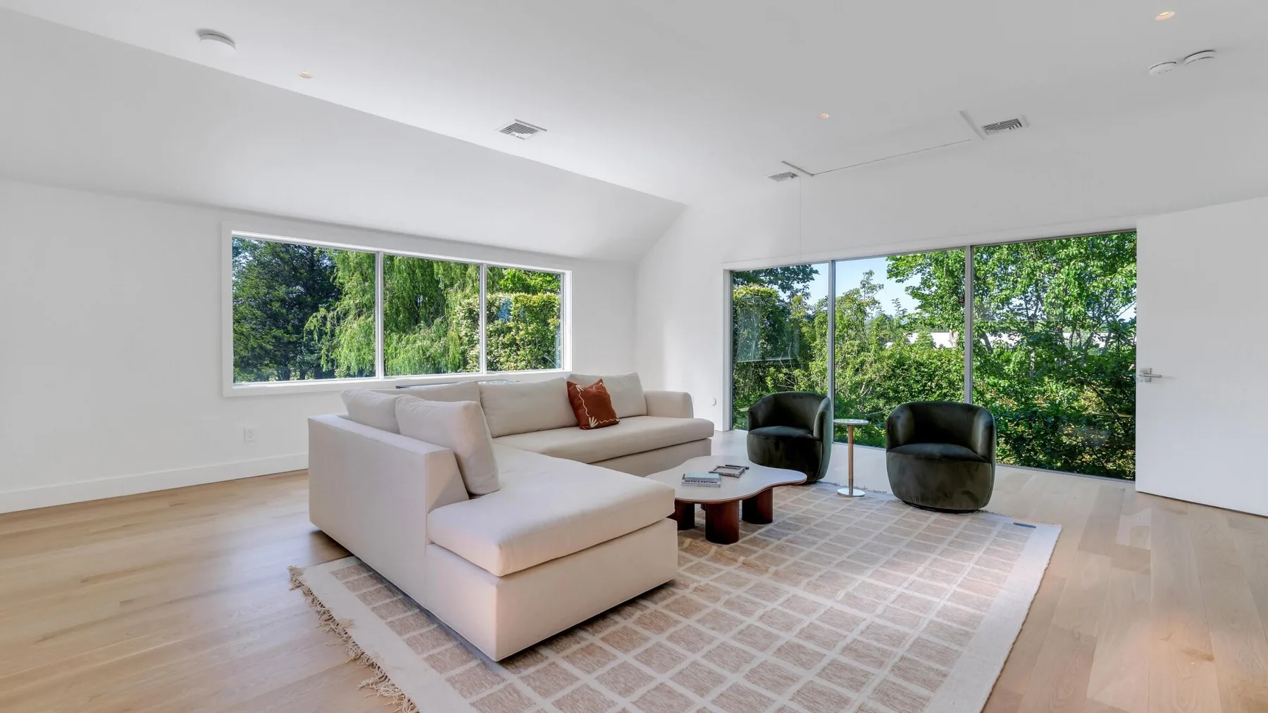 Una sala luminosa con un sofá seccional y grandes ventanas con vista a los árboles.