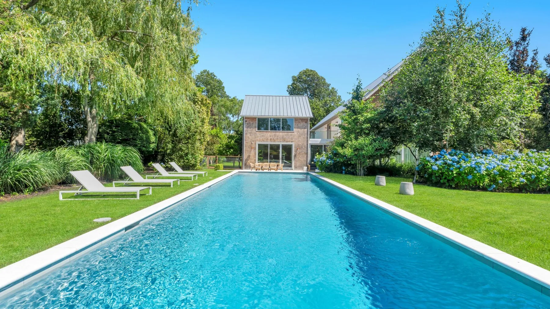 A long outdoor swimming pool stretches across a landscaped garden toward a modern house.