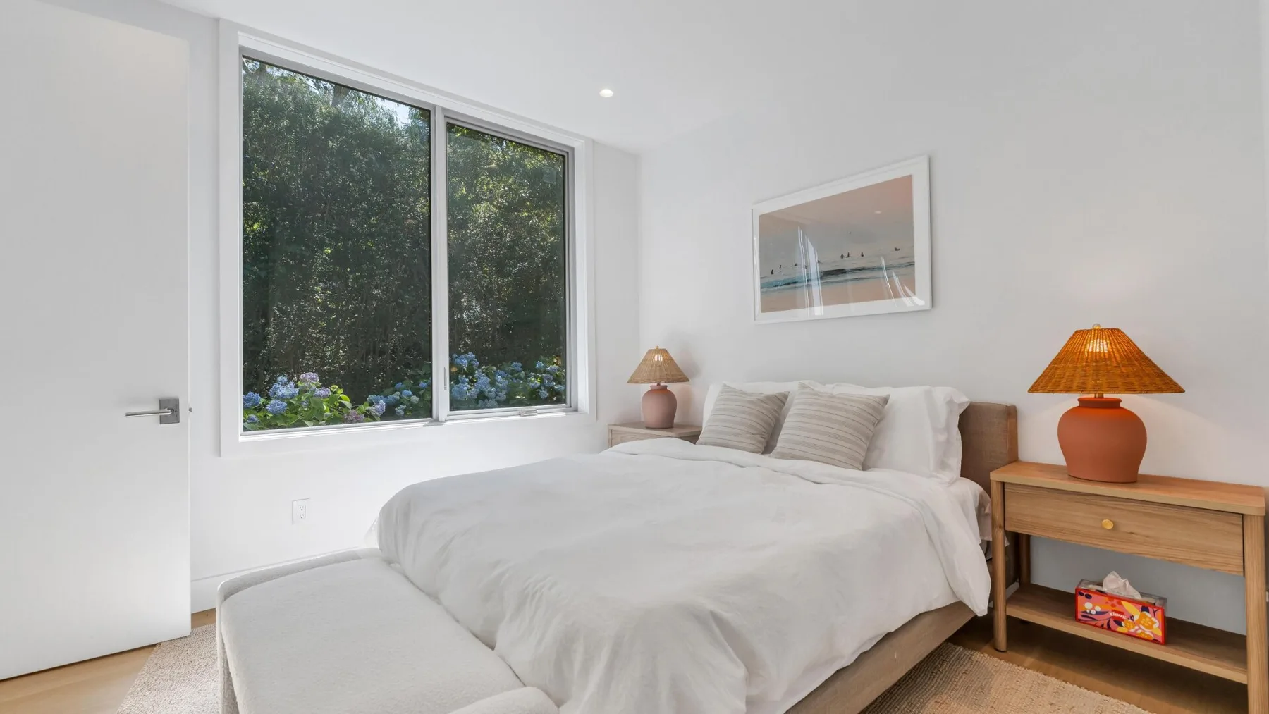 Un dormitorio luminoso con una cama grande junto a una ventana y una mesita de madera.