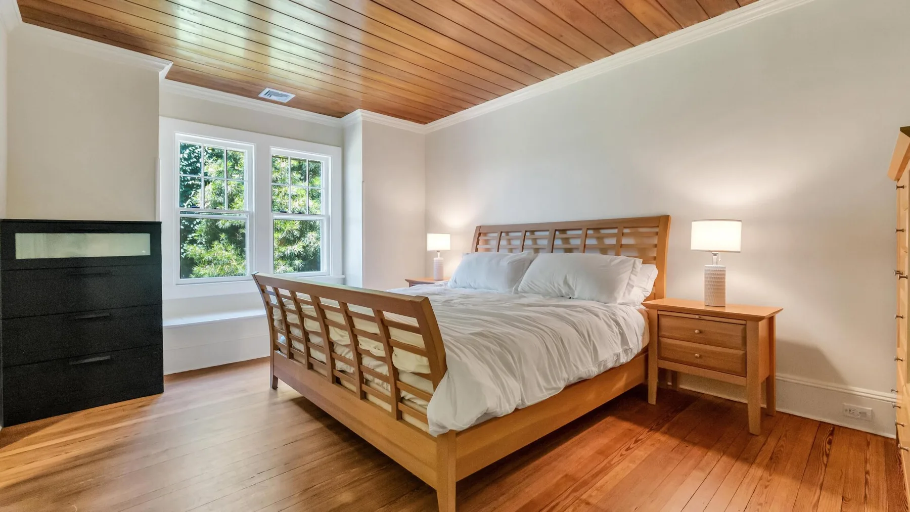 Un dormitorio con una cama de madera, mesitas de noche y ventanas grandes.