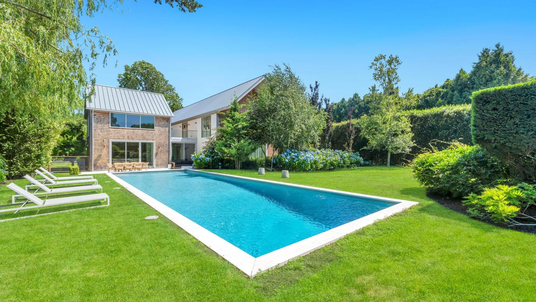 Una piscina junto a una casa moderna, rodeada de césped verde y árboles.