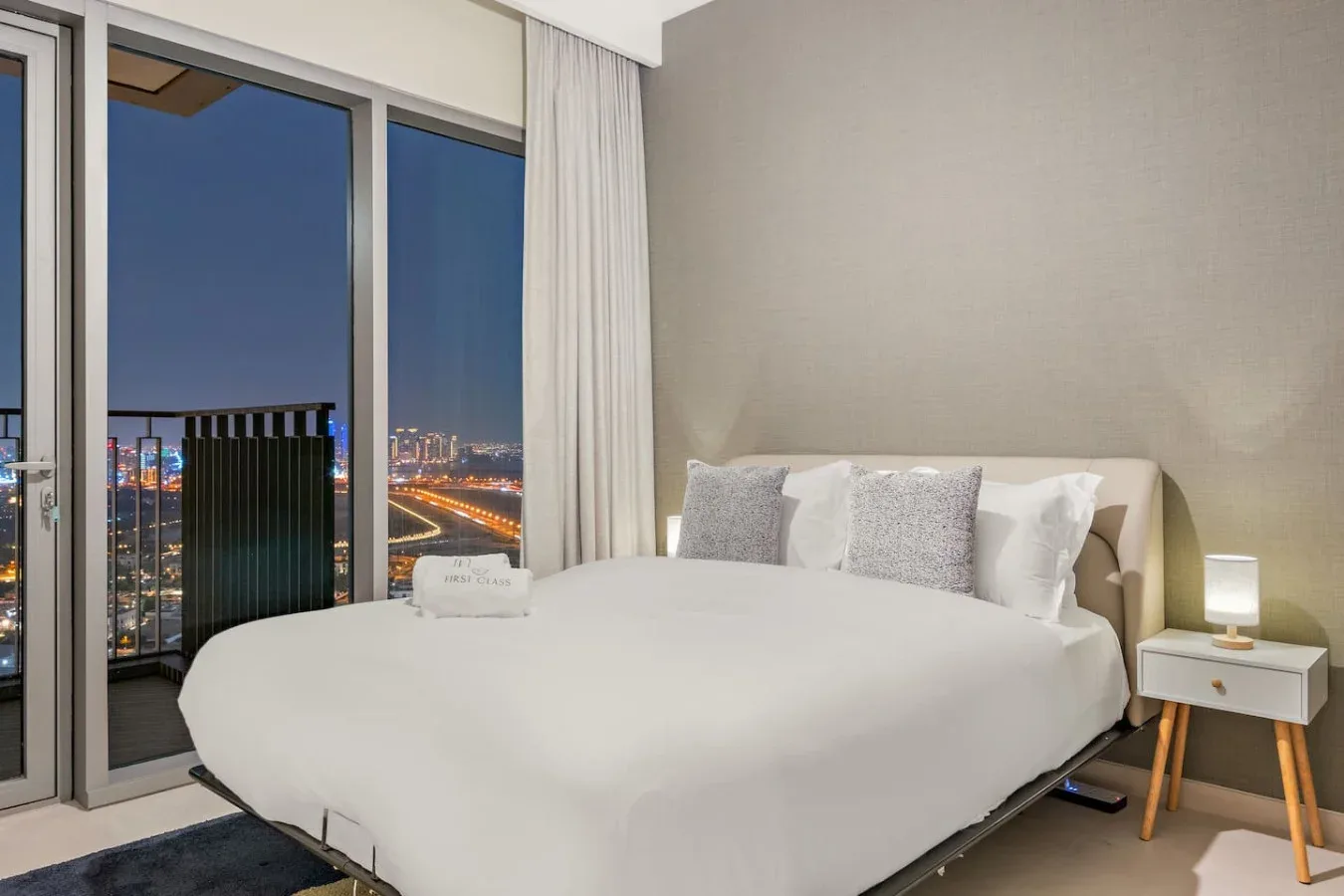 Un dormitorio con una cama tendida junto a grandes ventanas con vistas a las luces de la ciudad.