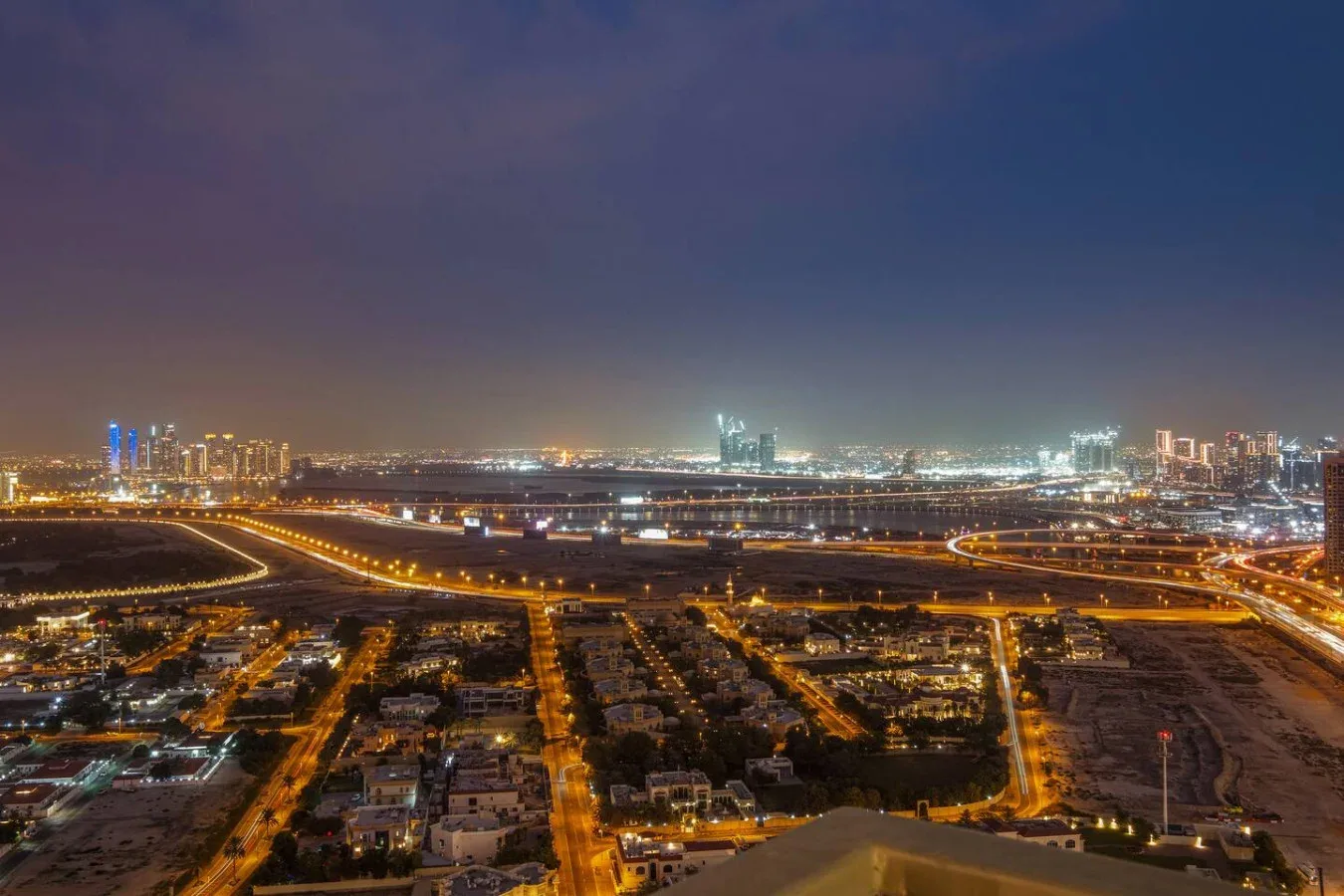 Un horizonte urbano nocturno brilla detrás de autopistas iluminadas y edificios bajos.