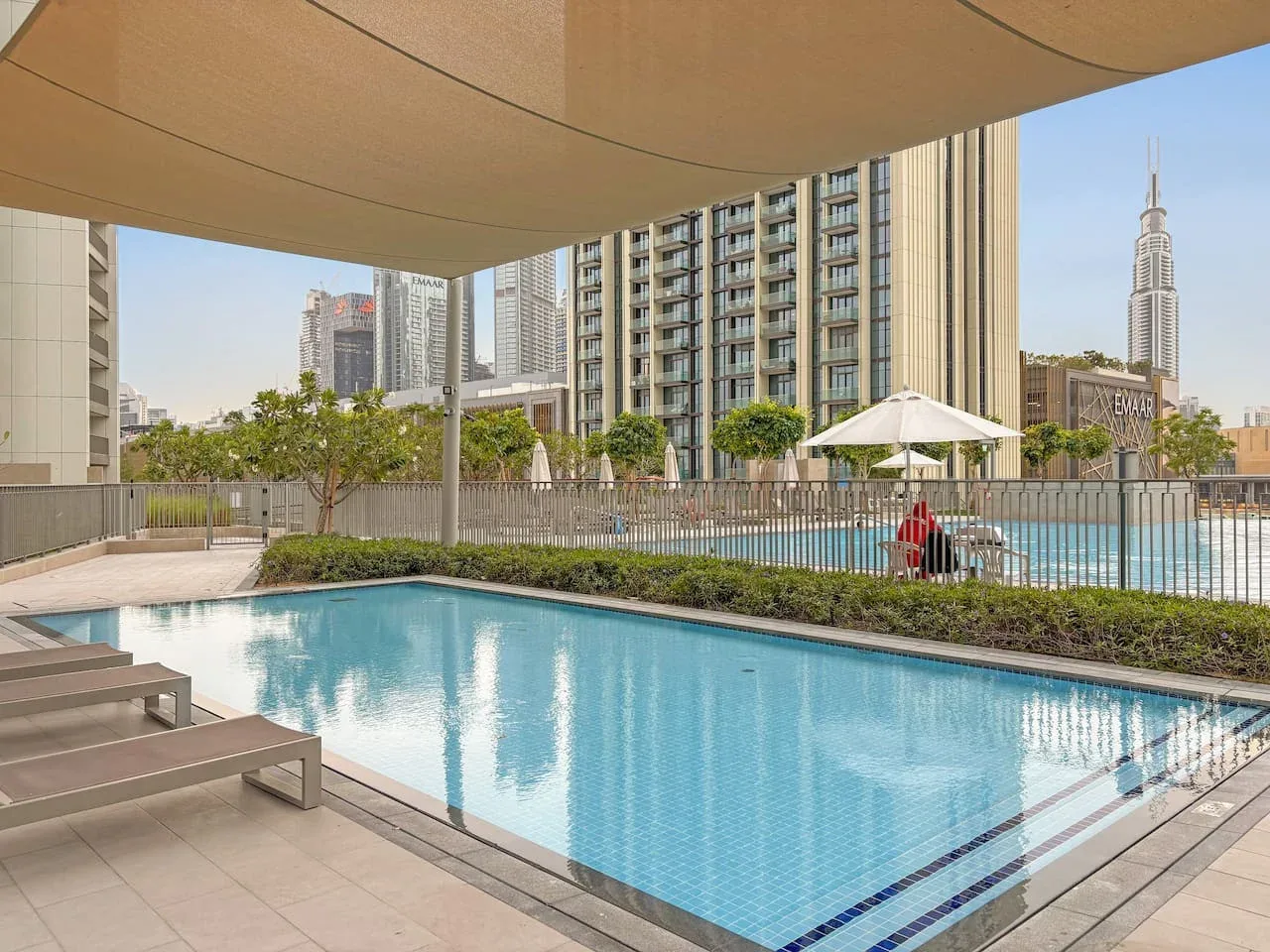 Una piscina al aire libre está bajo una cubierta de sombra, con edificios altos al fondo.