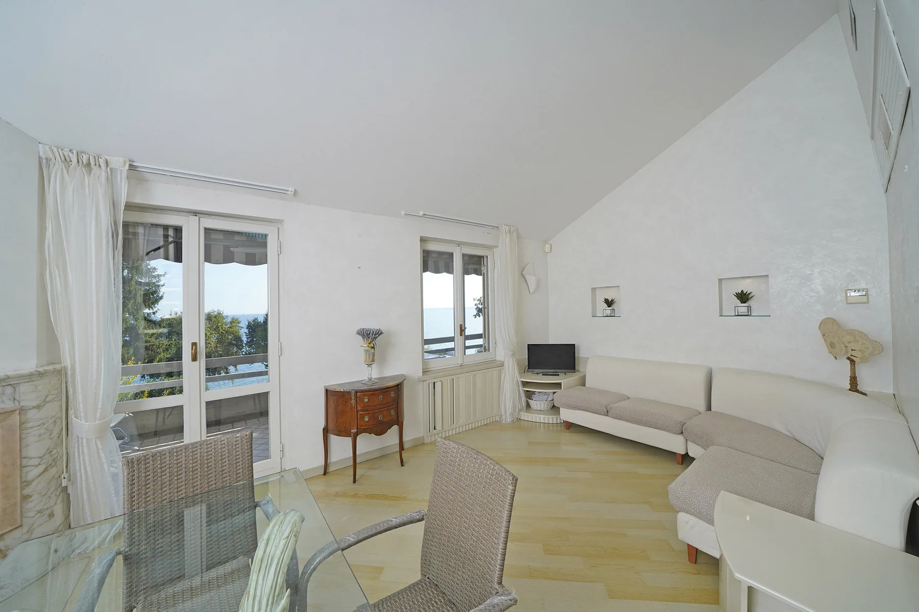 A bright living room with seating, a glass dining table, and windows opening onto a balcony.