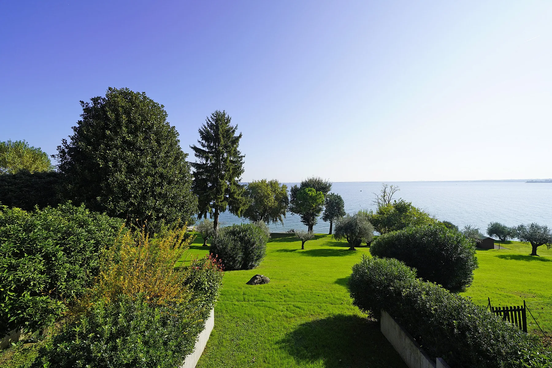 A green lawn with shrubs and trees slopes toward a calm waterfront.