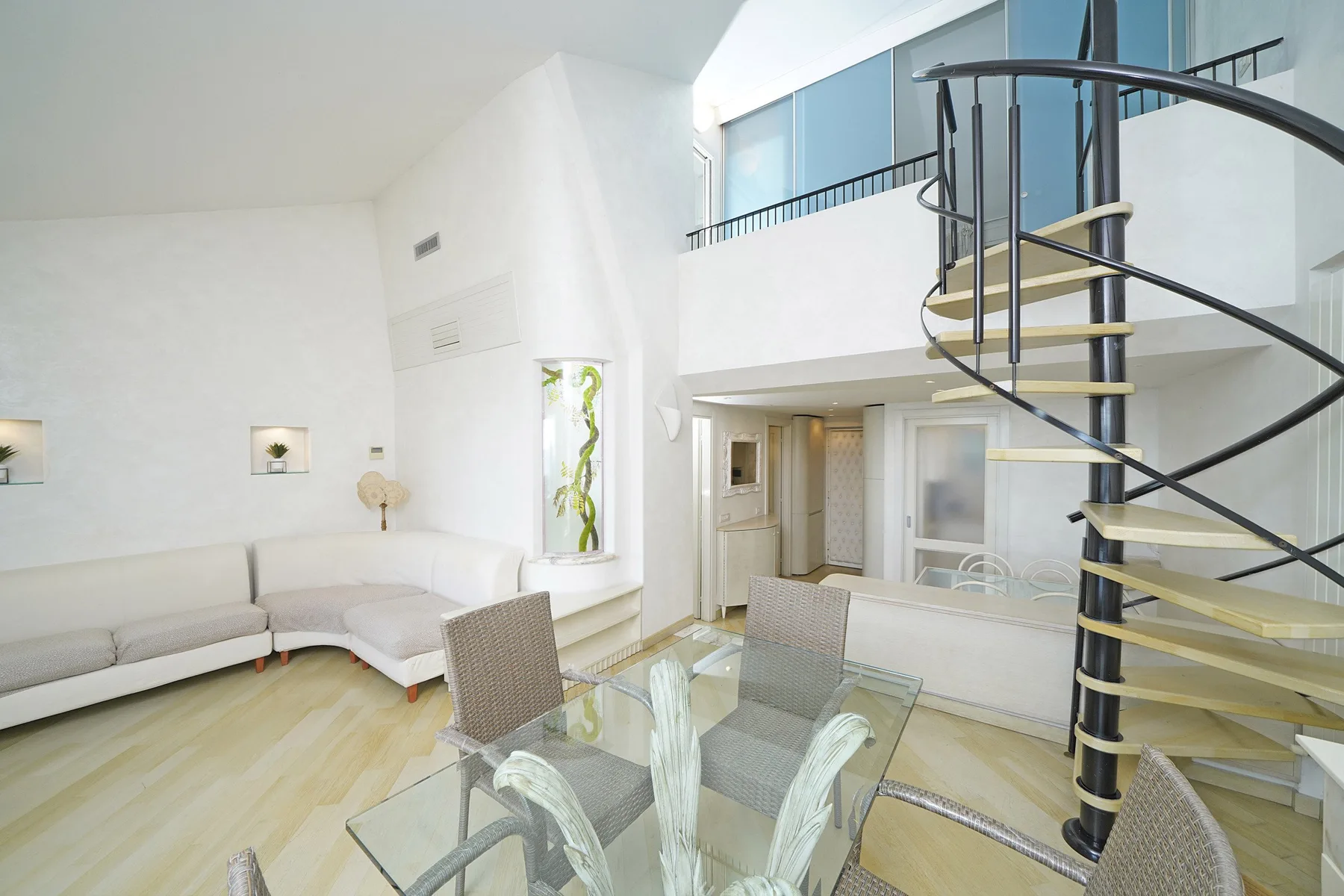 A bright living and dining area features a glass table and a curved staircase.