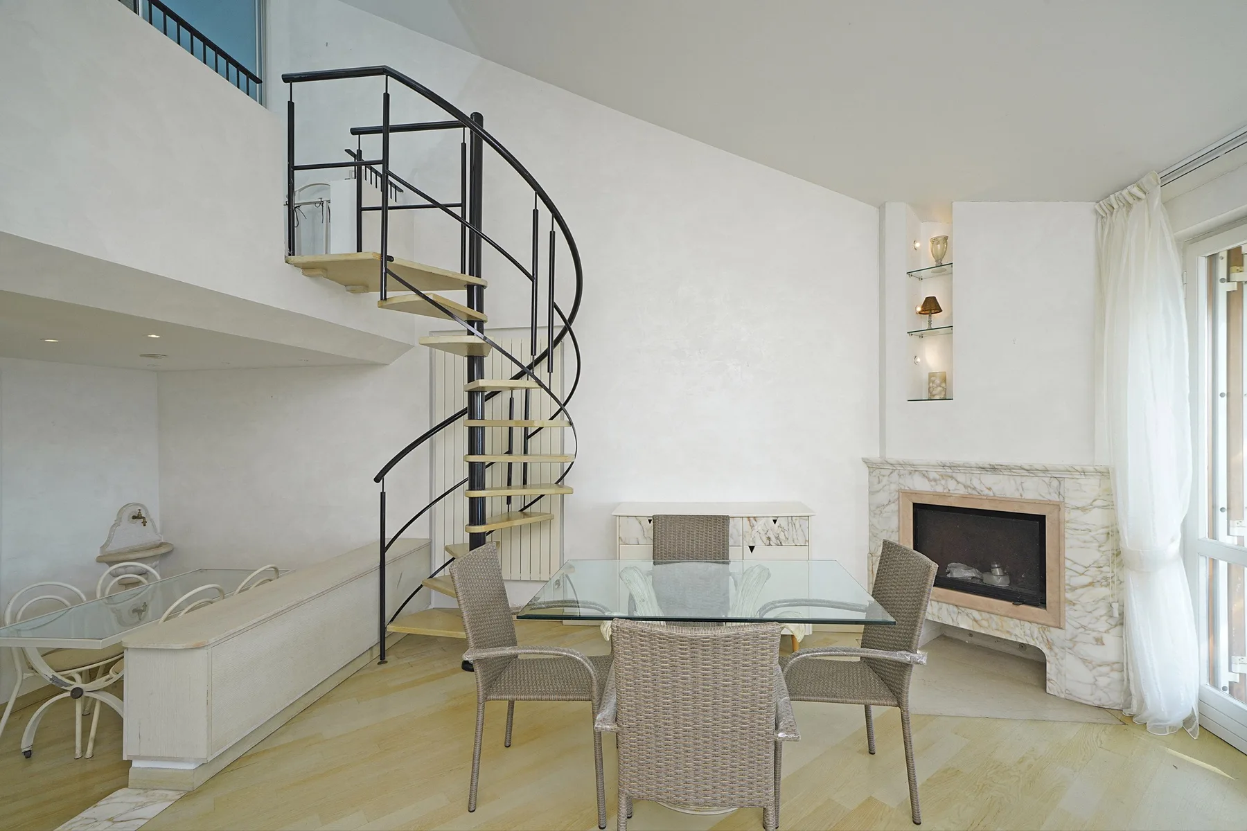 A bright living and dining area features a glass table and spiral staircase.