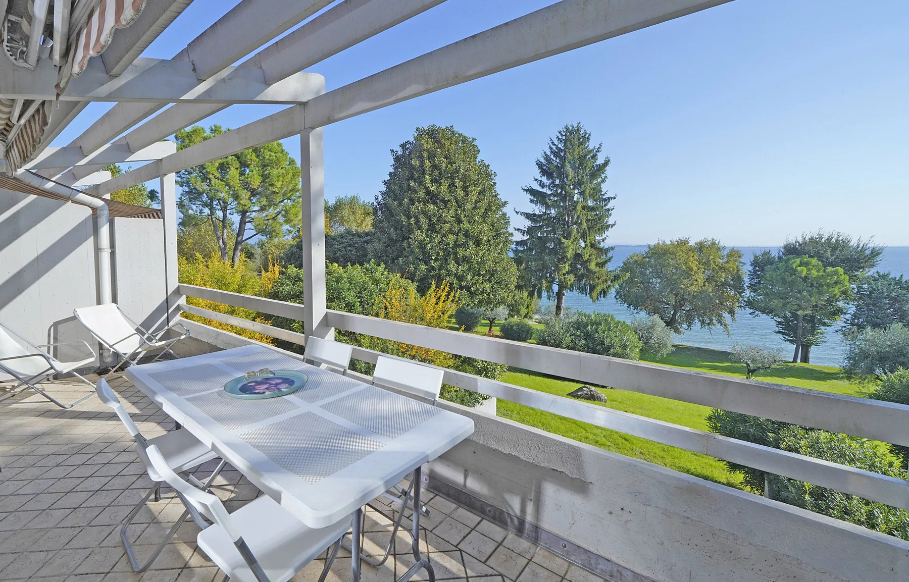 A furnished balcony overlooks a green lawn, trees, and a body of water.