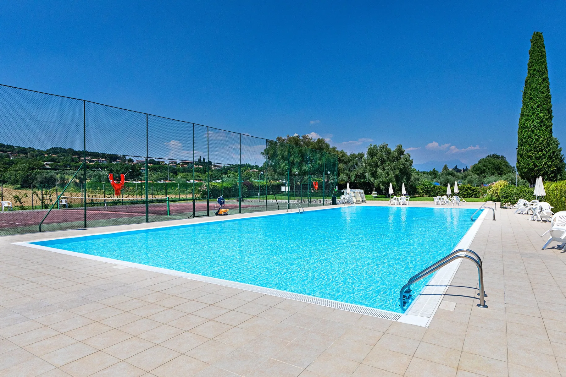 An outdoor swimming pool beside a fenced tennis court under a clear sky.