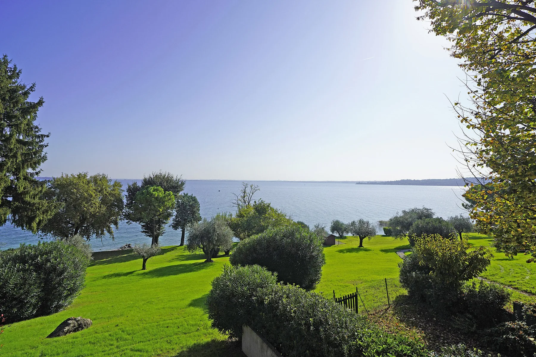 A green lawn with shrubs overlooks a calm body of water beneath a clear sky.