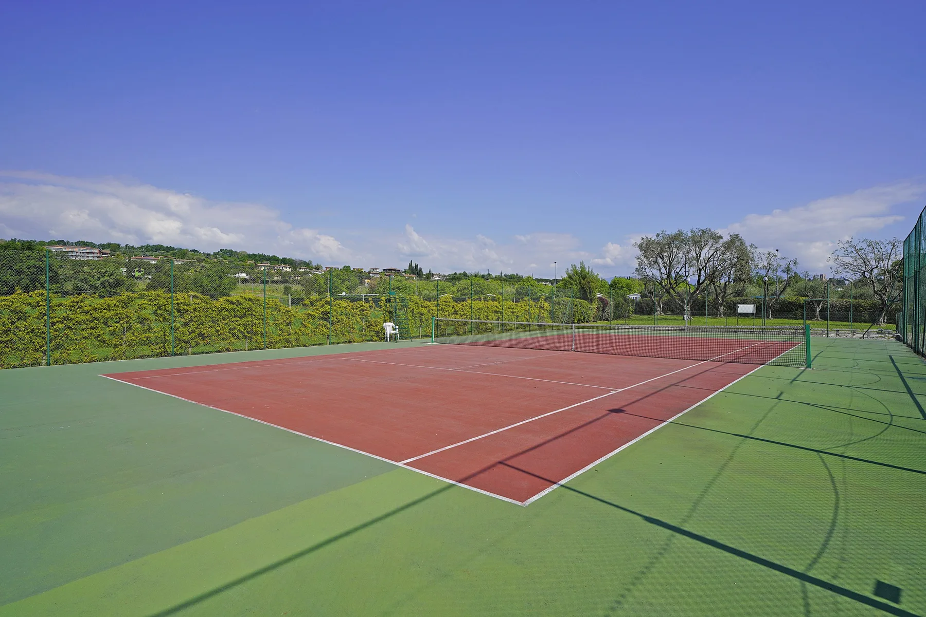 An outdoor tennis court with a red playing surface and green surrounding area.