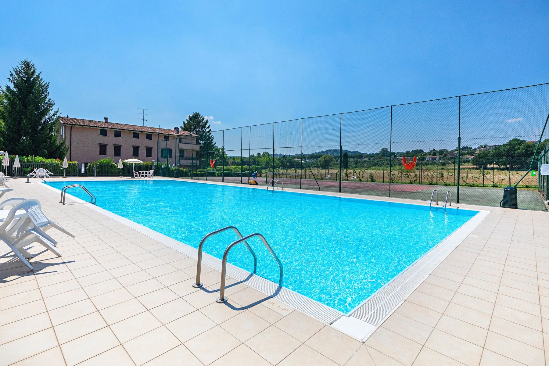 An outdoor swimming pool sits beside a fenced sports court under a clear sky.
