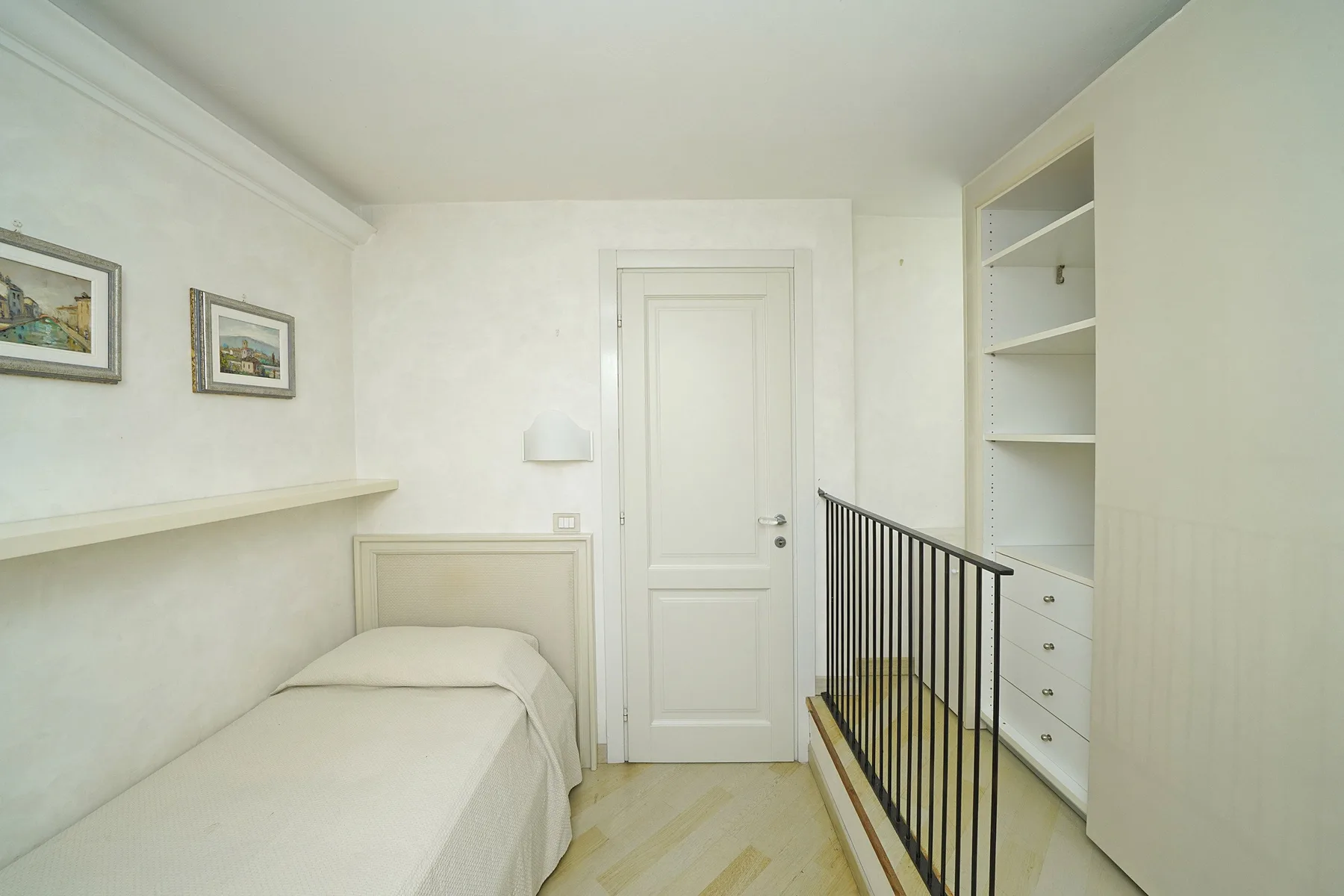 A bright bedroom with a single bed beside built-in shelving and a railing.