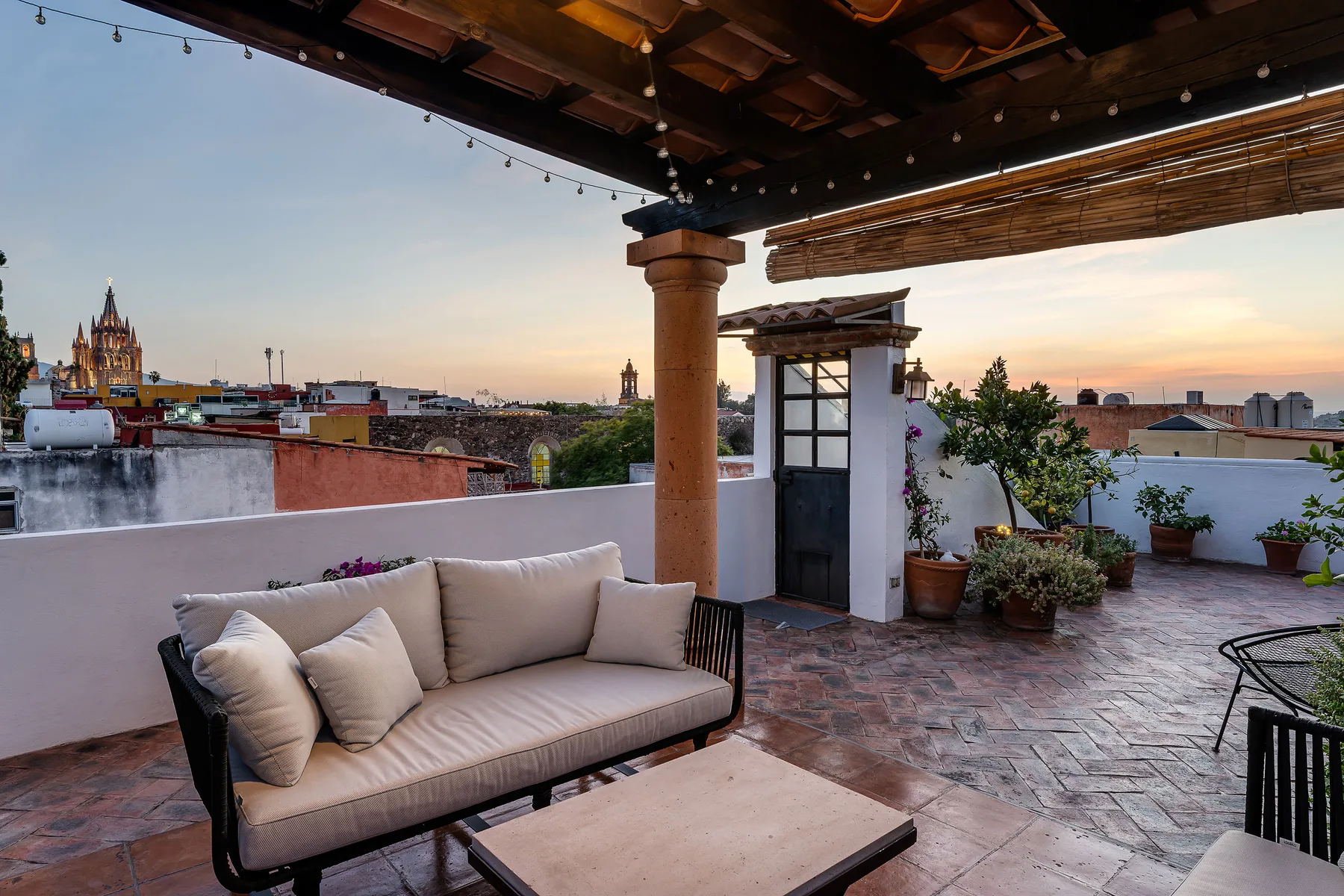 A rooftop terrace has cushioned seating, potted plants, and views of the surrounding city.