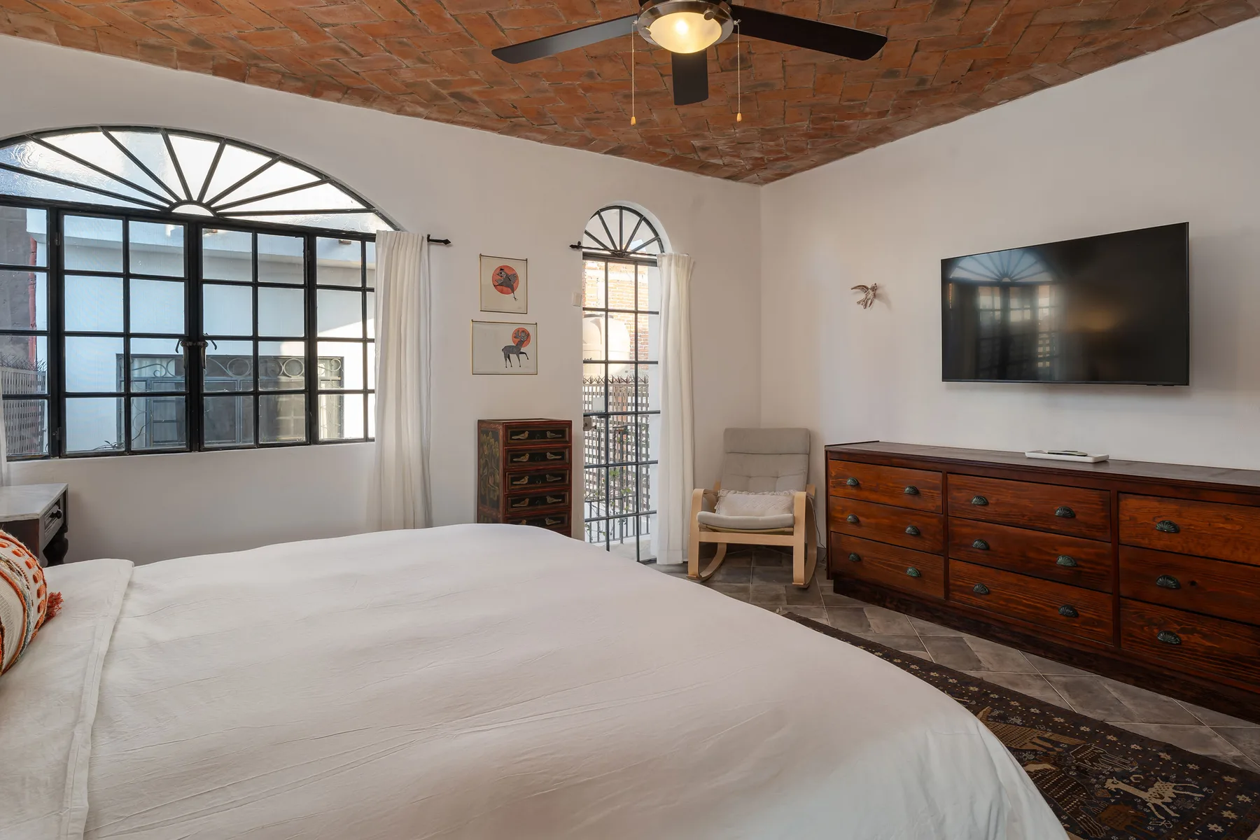 A bedroom with a large bed, arched windows, and a wooden dresser.