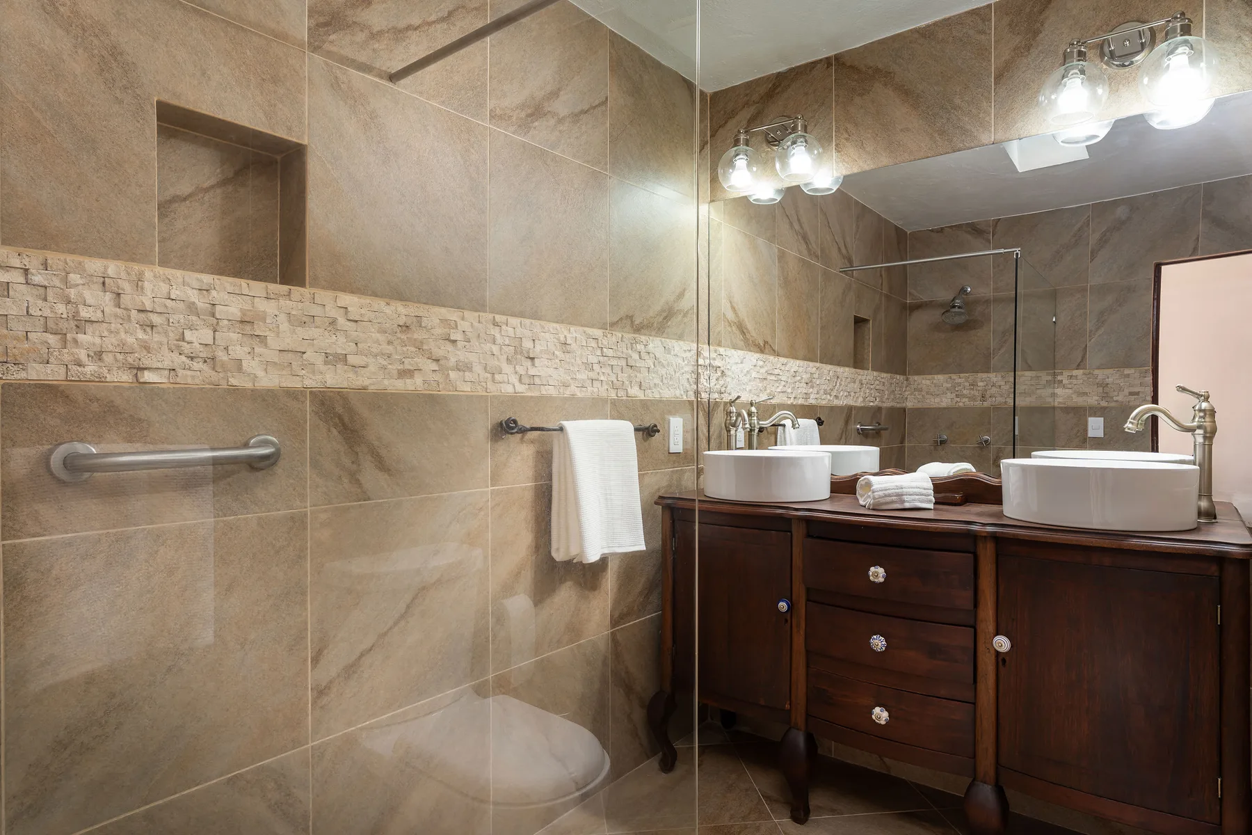 A tiled bathroom with a vanity, sinks, and a glass shower.