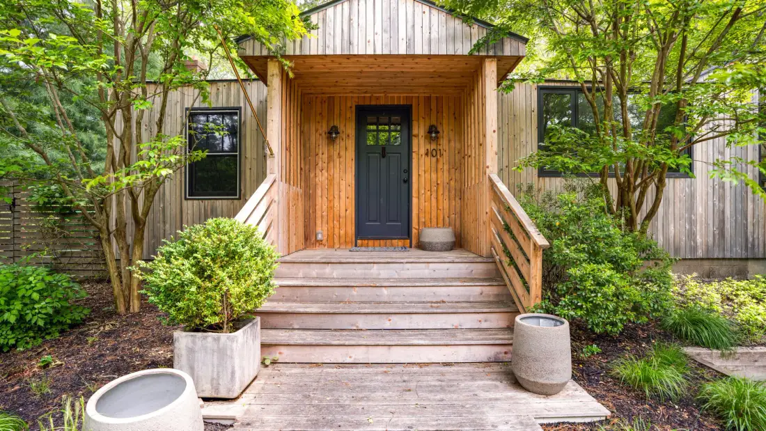 A wooden building entrance with steps, a dark door, and surrounding greenery.
