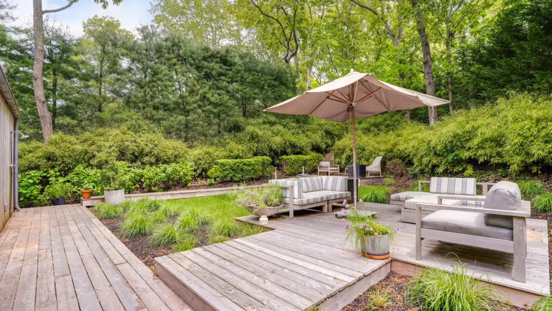 An outdoor deck with seating beneath a large umbrella, surrounded by greenery.