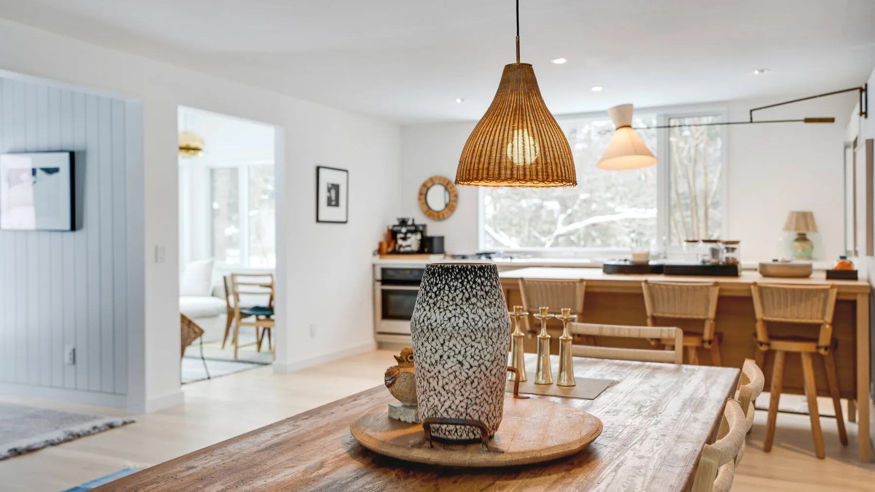 A bright dining area has a long wooden table, woven pendant light, and kitchen island.