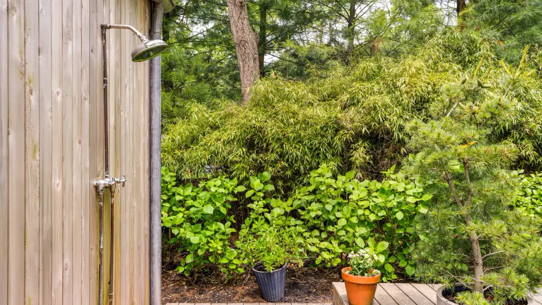An outdoor shower beside a leafy garden with potted plants.
