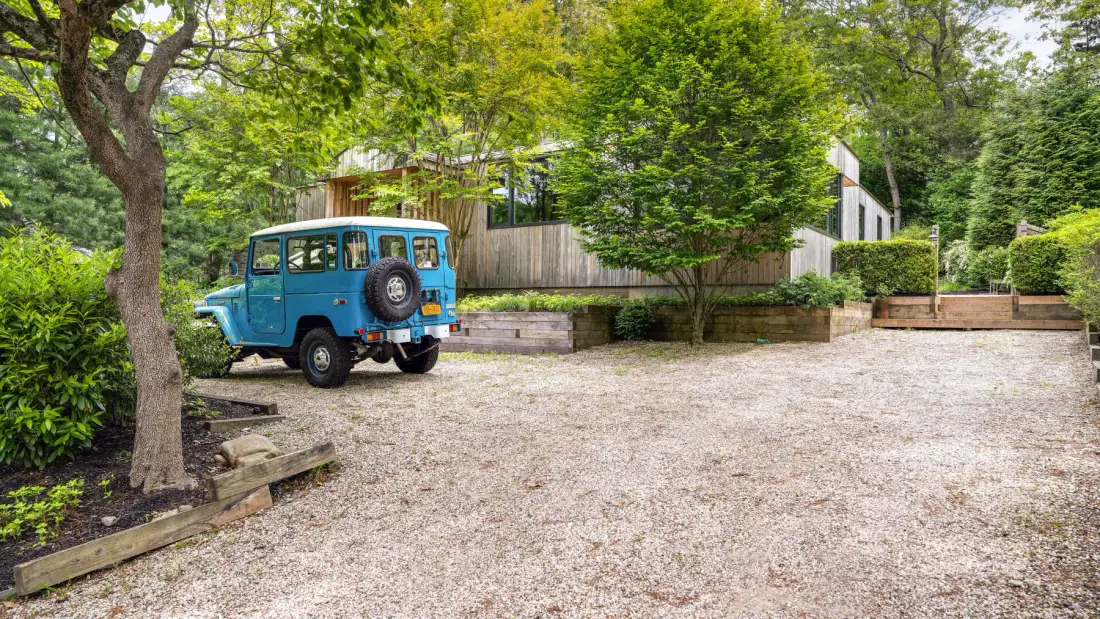 A gravel driveway bordered by trees leads toward a modern building, with a blue vehicle parked nearby.