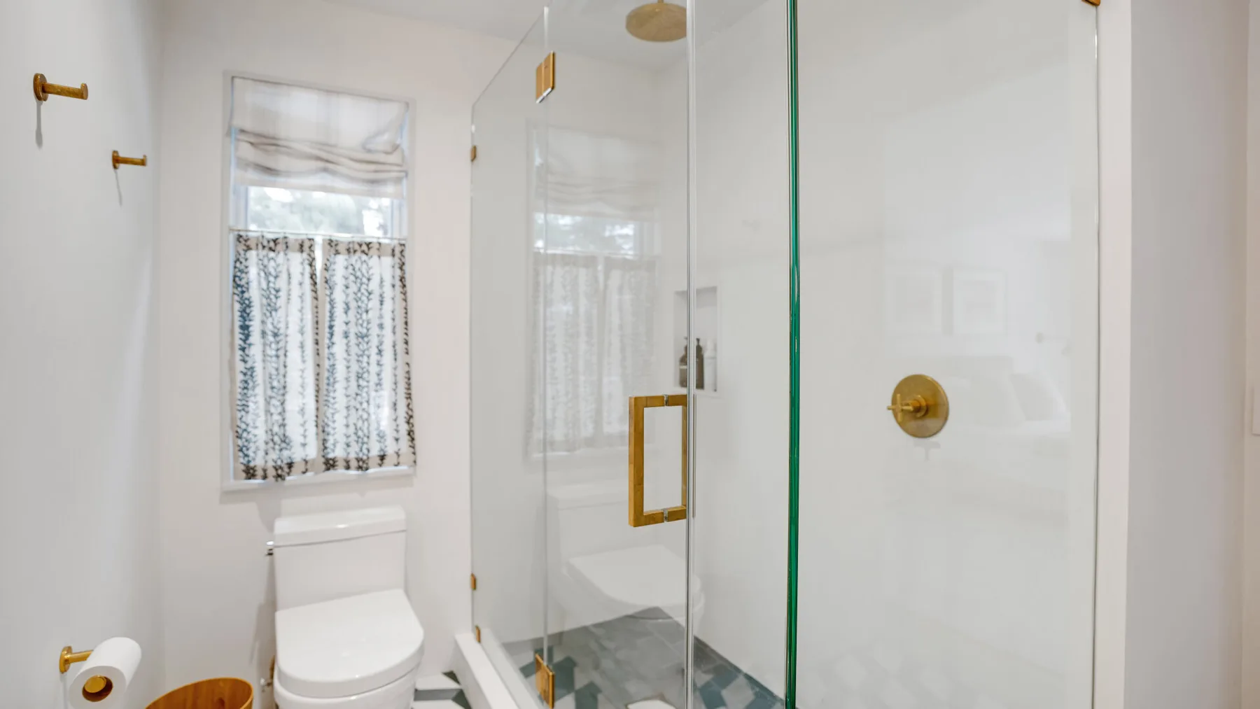 A bright bathroom with a glass shower and a toilet beside a curtained window.