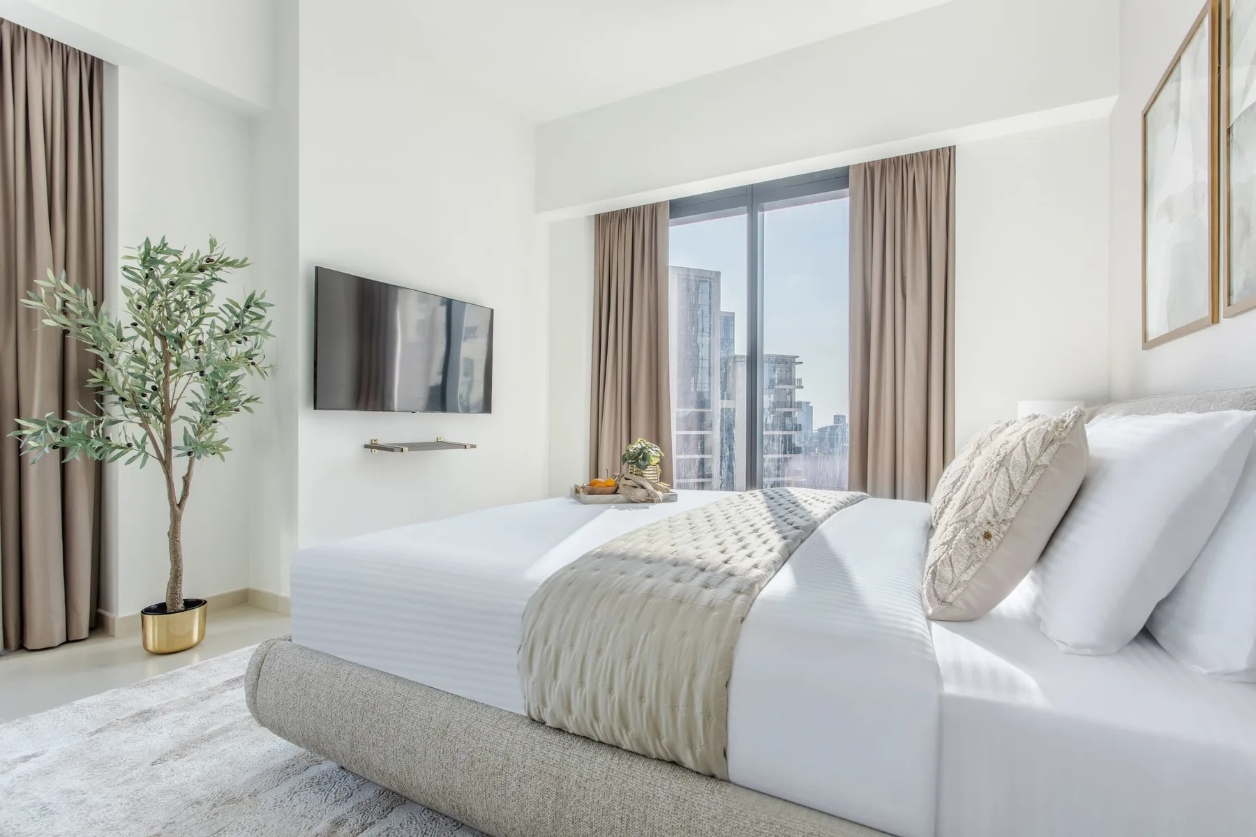 Un dormitorio luminoso con una cama tendida y una gran ventana con vista a edificios de la ciudad.