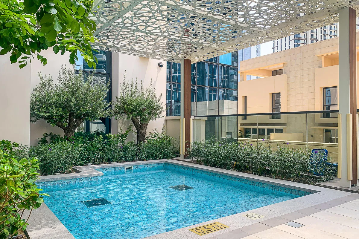 Una piscina está bajo una cubierta calada, rodeada de vegetación y edificios modernos.