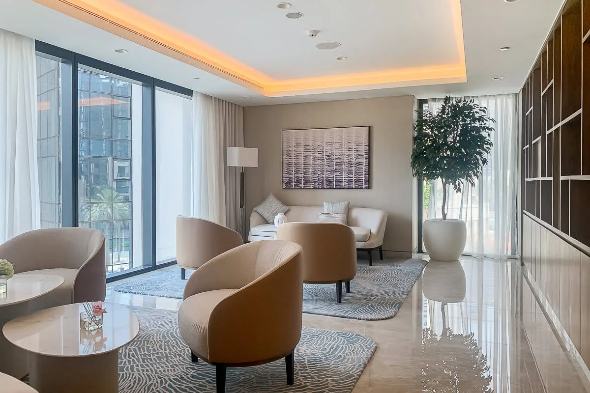 Una sala luminosa con asientos, grandes ventanales y un árbol en maceta.