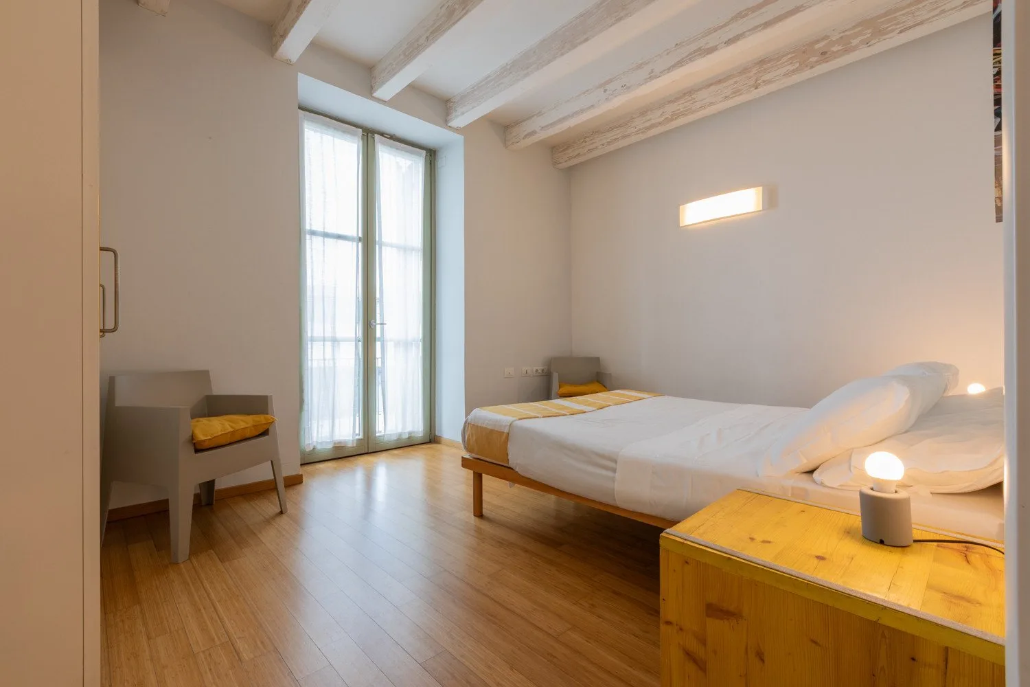 Un dormitorio luminoso con una cama, suelo de madera y ventanas altas.