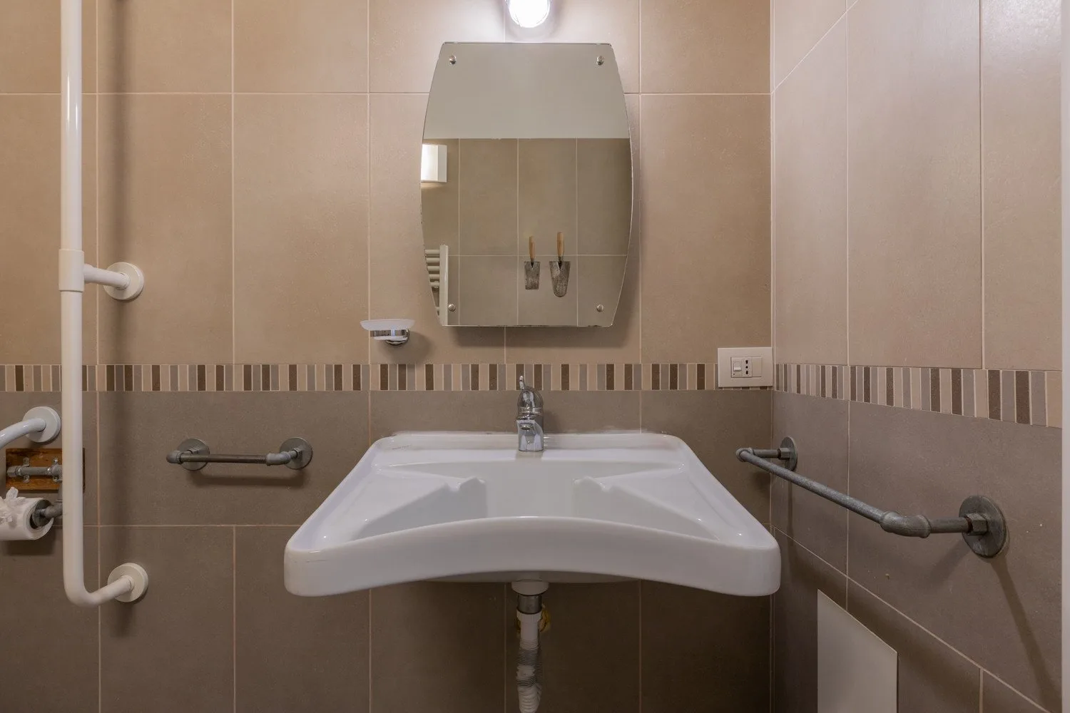 Un baño con azulejos, lavabo de pared, espejo y barras de apoyo.