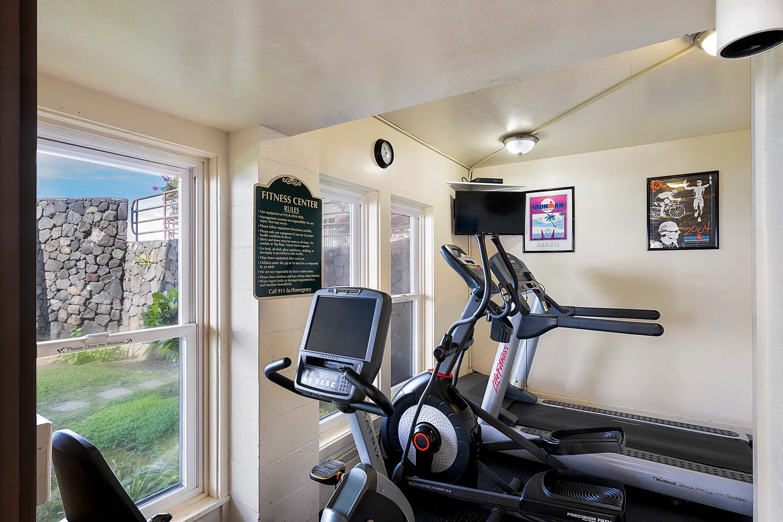 Un gimnasio con equipos de ejercicio y ventanas con vista a un muro de piedra.