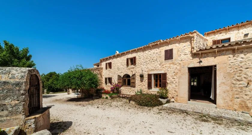 A stone building faces a sunlit gravel courtyard under a clear blue sky.
