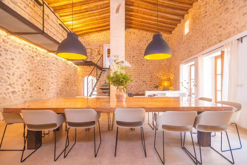 A long dining table sits beneath pendant lights in a bright stone-walled room.