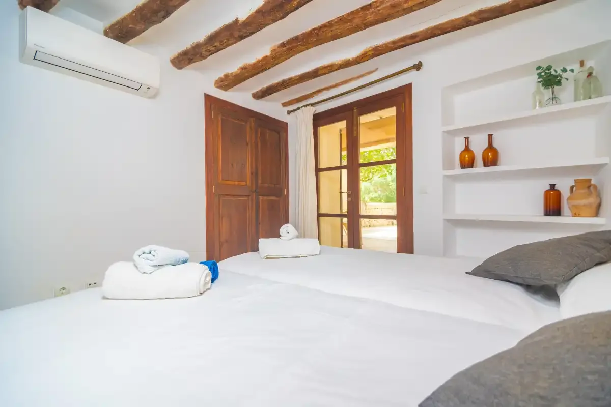 A bright bedroom with white bedding, wooden ceiling beams, and built-in shelves.