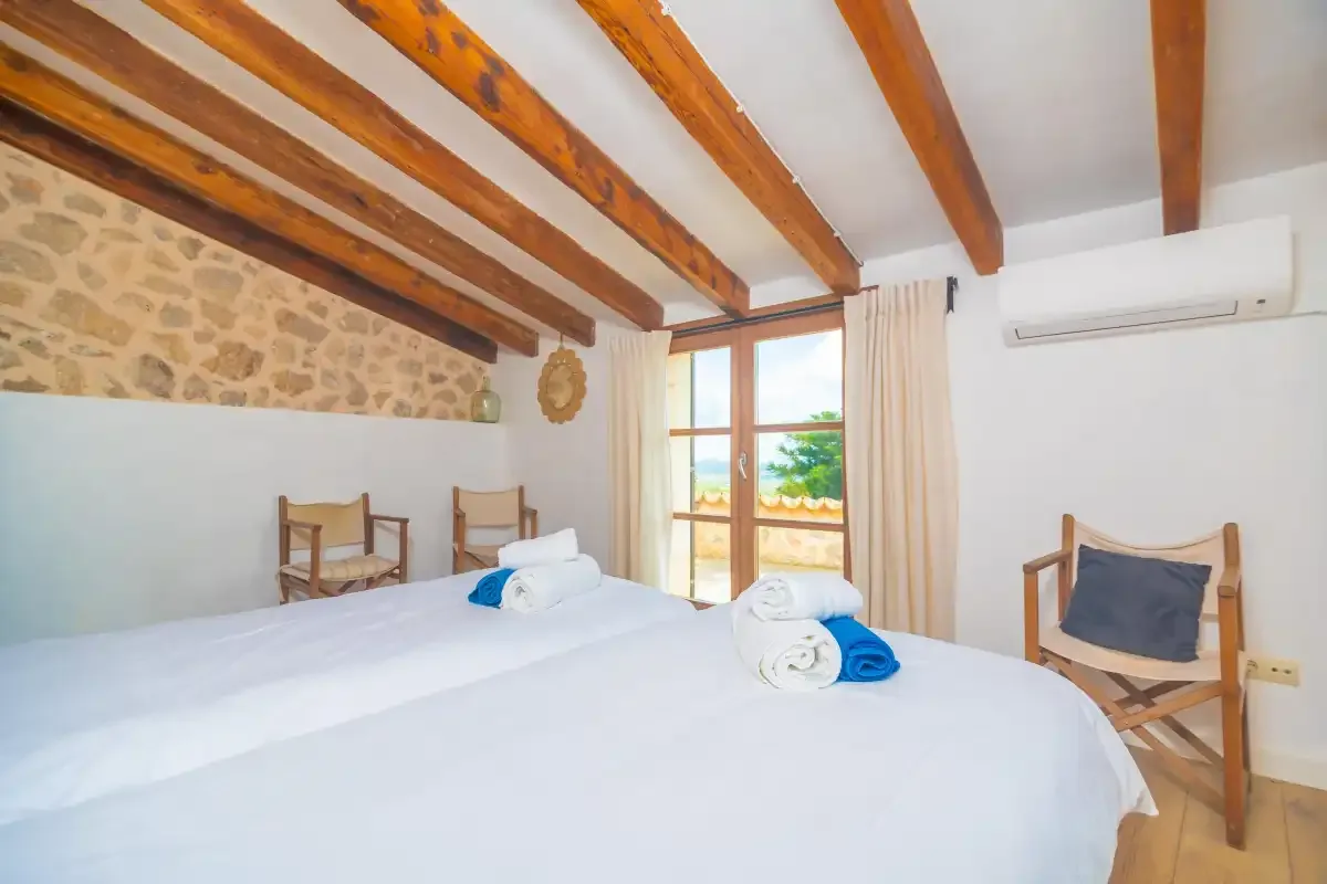 A bright bedroom with white bedding, wooden chairs, and exposed ceiling beams.