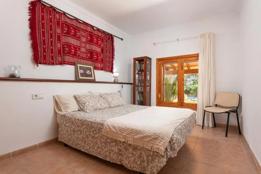 A bedroom with a bed beneath a red patterned wall hanging and beside a wooden window.