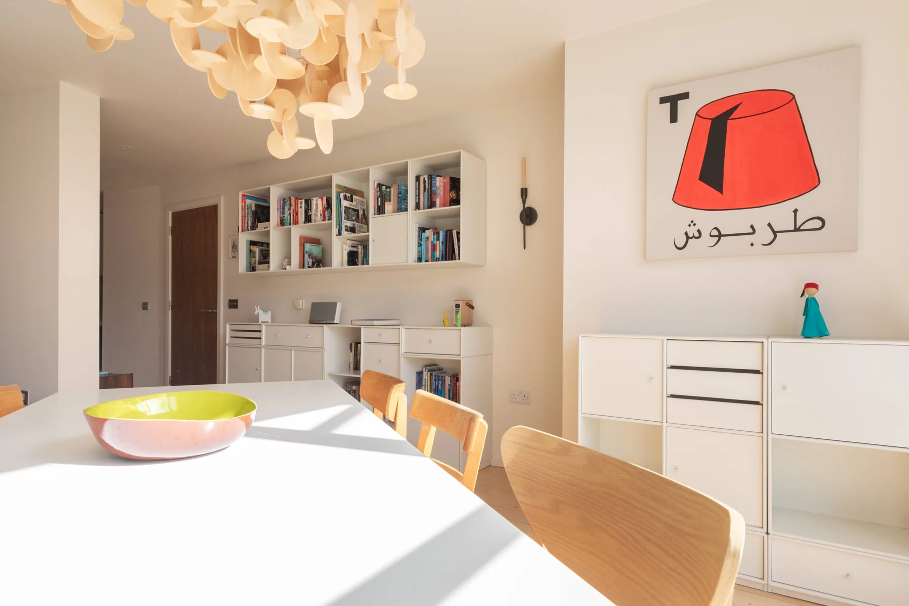 Un comedor luminoso con una mesa larga, asientos de madera y estantes abiertos en la cocina.