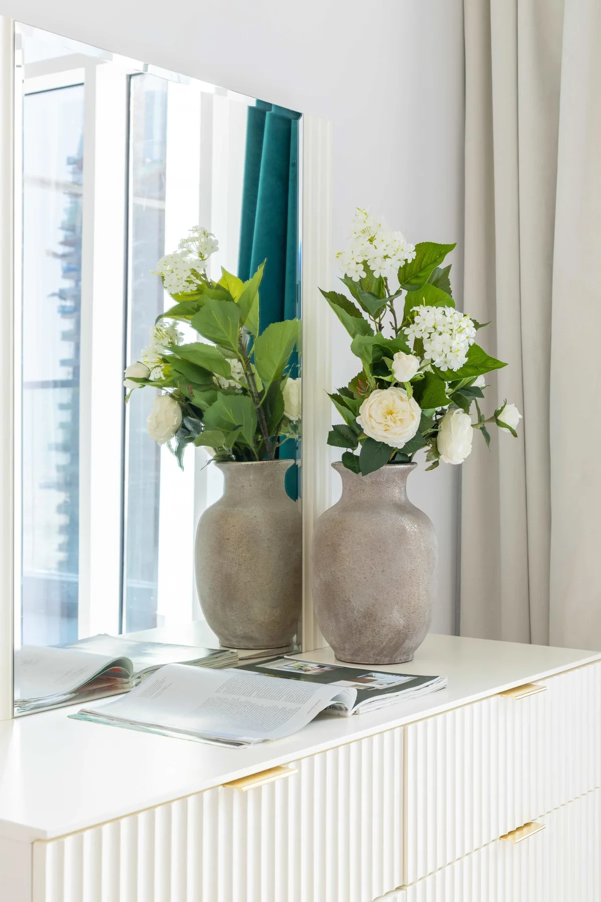 A white console table displays a vase of white flowers beside a tall mirror.