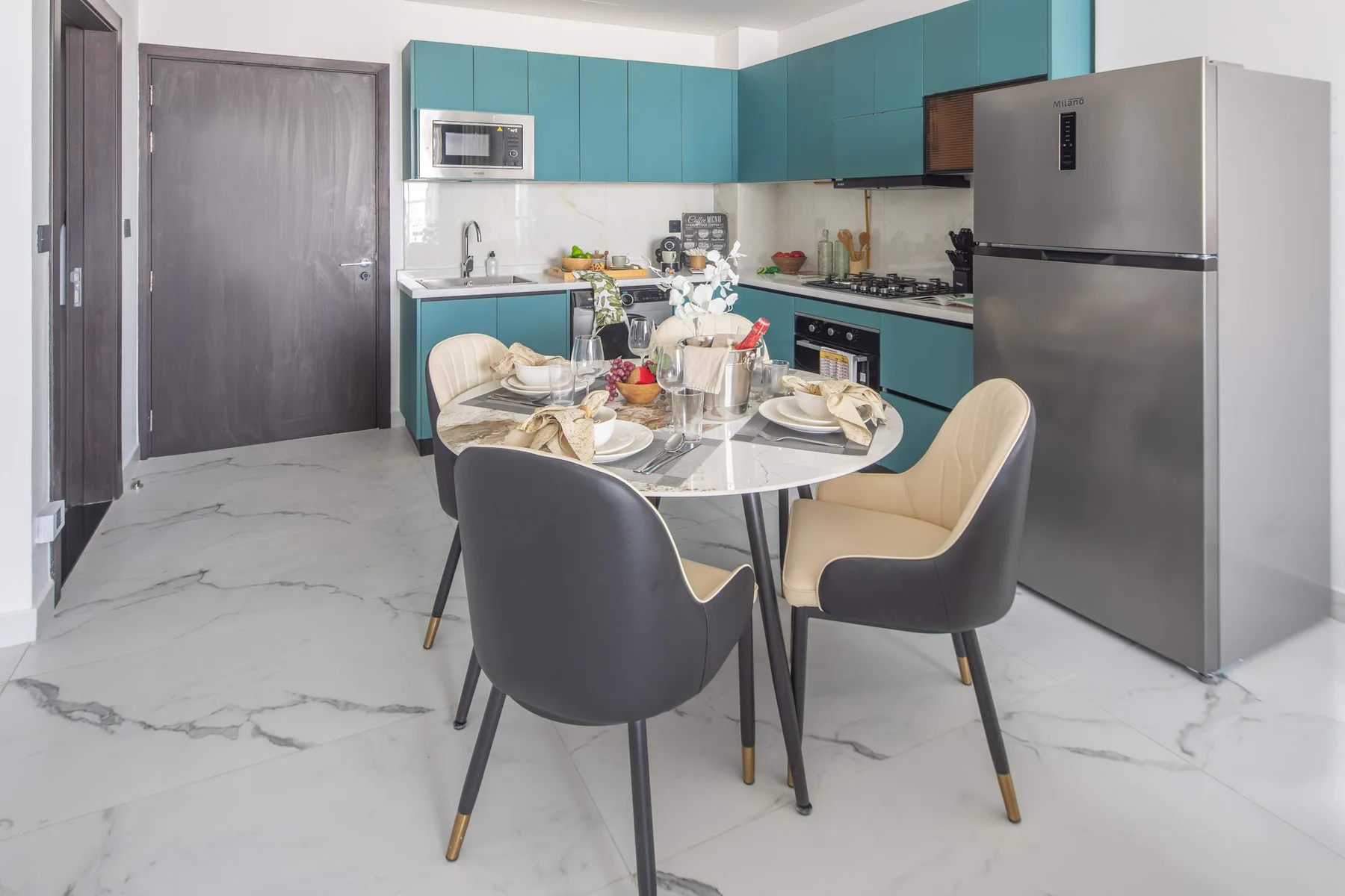 A dining area with a set table sits beside a teal kitchen and stainless-steel refrigerator.
