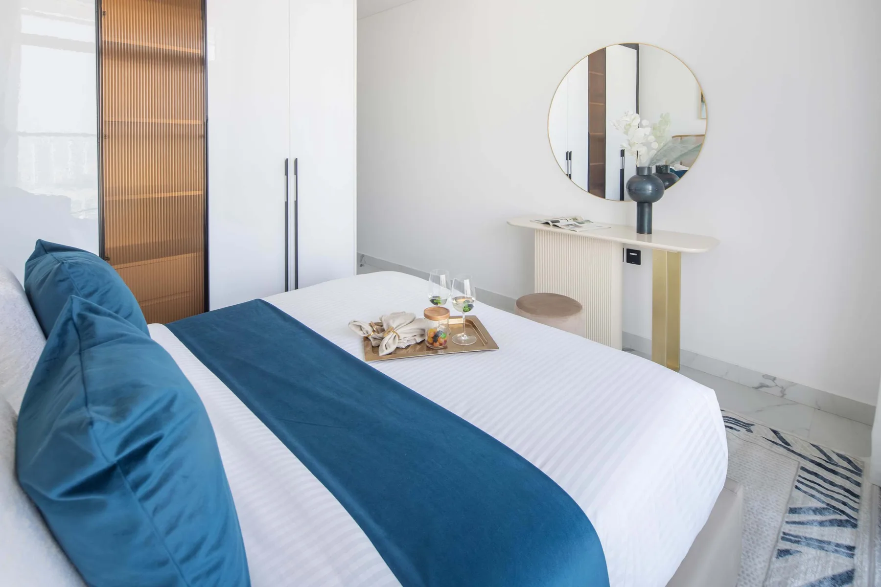 A bedroom with a white bed, blue bedding, and a round wall mirror.