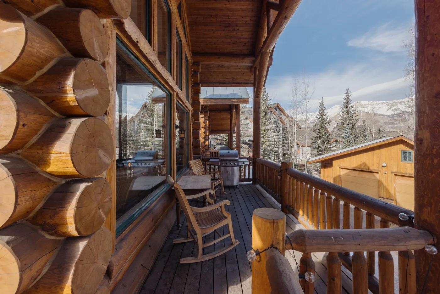 Un porche de cabaña de madera con mecedoras mira hacia montañas nevadas y árboles.