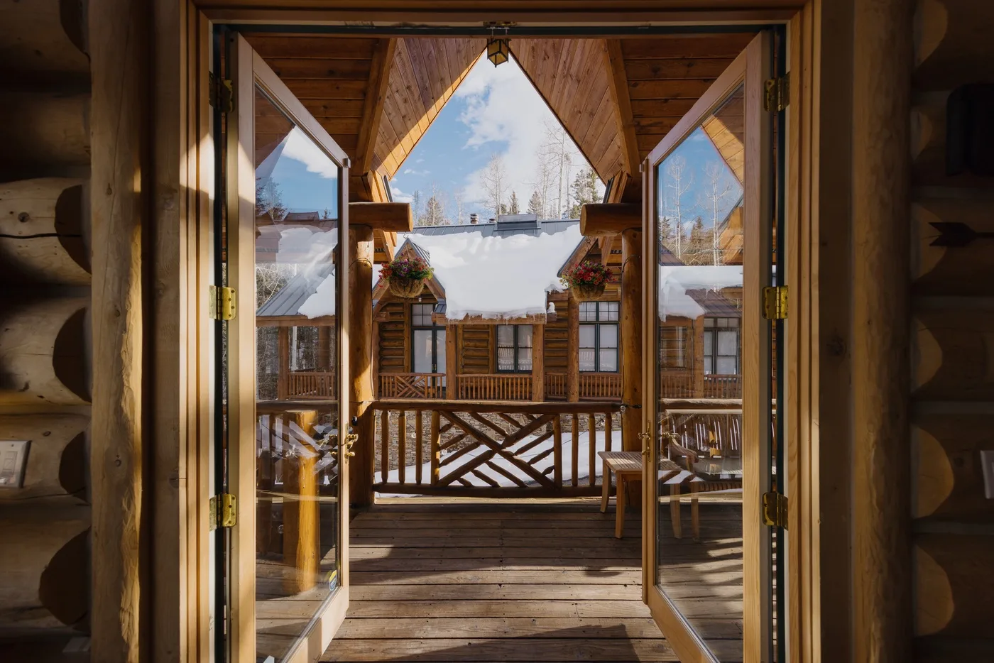 Un porche de madera enmarca la vista de una cabaña nevada bajo un cielo azul.