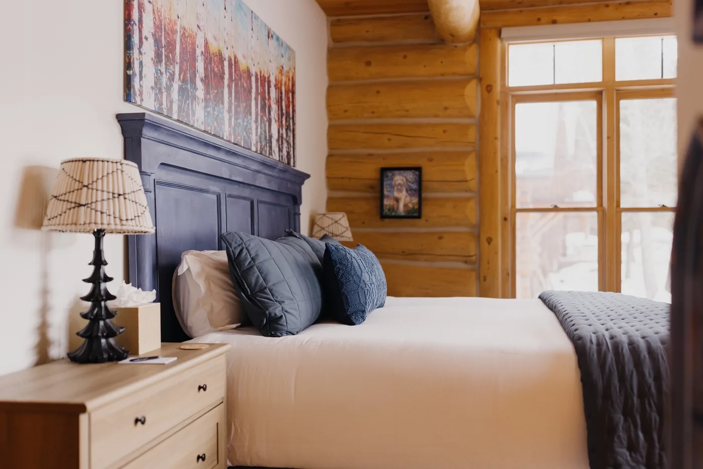 Dormitorio con una cama, un cabecero oscuro y grandes ventanas junto a una pared revestida de madera.
